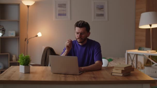 Um homem em uma camiseta roxa senta-se em uma mesa em seu quarto e trabalha em um laptop. Um homem sentado em um computador esfrega seu queixo e pensa como resolver problemas com uma tarefa de estudo. Imagens 4k de alta qualidade - Filmagem, Vídeo