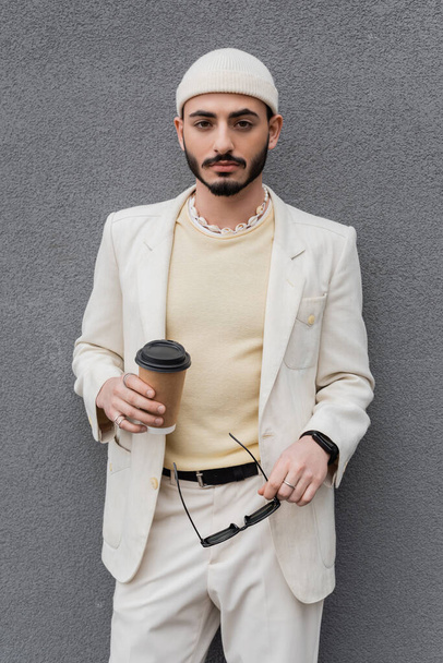 Fashionable gay man in suit holding coffee to go and sunglasses near wall outdoors  - Foto, imagen