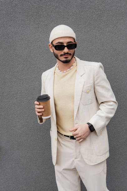 Modieuze gay man in beige pak en zonnebril houden papier beker buiten  - Foto, afbeelding