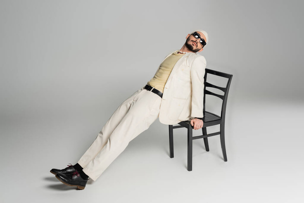 Well dressed gay man in sunglasses posing with chair on grey background  - Photo, Image