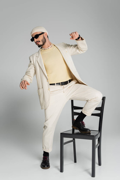 Trendy gay man in suit and sunglasses posing near chair on grey background  - Fotó, kép