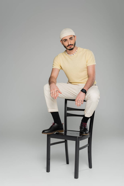 Bearded gay man in hat sitting on chair on grey background  - Zdjęcie, obraz