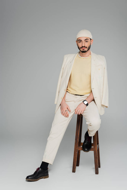 Fashionable gay man in beige suit and hat sitting on chair on grey background  - Foto, immagini