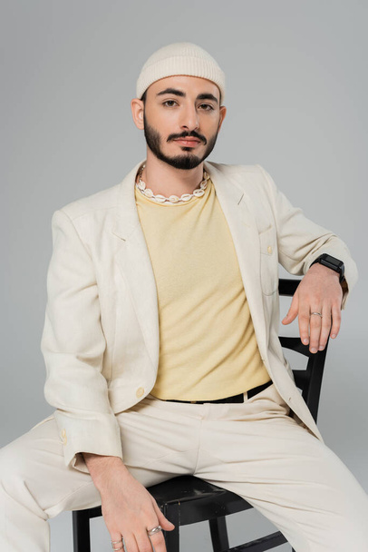 Bearded homosexual man in suit ad seashell necklace sitting on chair isolated on grey   - Foto, Imagem
