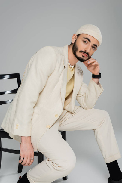 Trendy homosexual man in hat and beige suit posing on chair on grey background  - Photo, Image