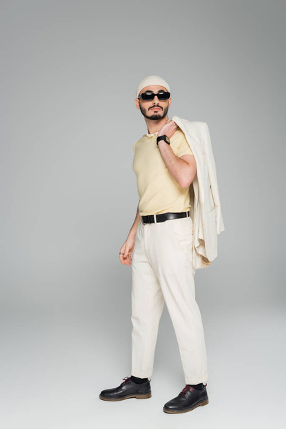 Stylish gay man in sunglasses holding jacket while standing on grey background  - Photo, Image