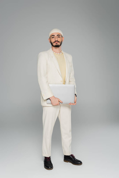 Stylish gay man in beige suit holding laptop on grey background  - Photo, Image