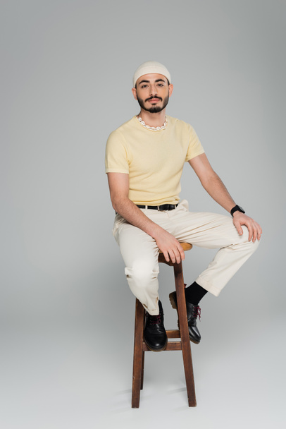Full length of trendy homosexual man sitting on chair on grey background  - Foto, Imagen