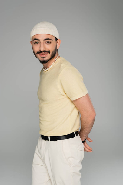 Portrait d'homme homosexuel branché en chapeau et collier coquillage souriant à la caméra isolé sur gris   - Photo, image