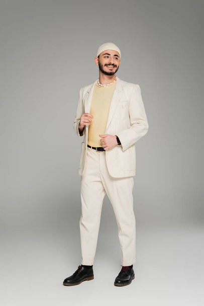 Cheerful homosexual man in beige suit and hat touching jacket on grey background  - Фото, изображение