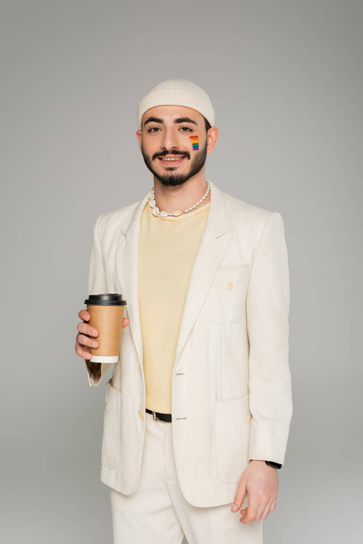 Fashionable gay man with lgbt flag on cheek holding coffee to go isolated on grey   - Photo, Image
