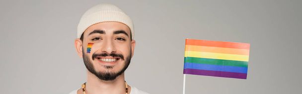 alegre homosexual hombre en sombrero mirando cámara cerca lgbt bandera aislado en gris, bandera  - Foto, Imagen