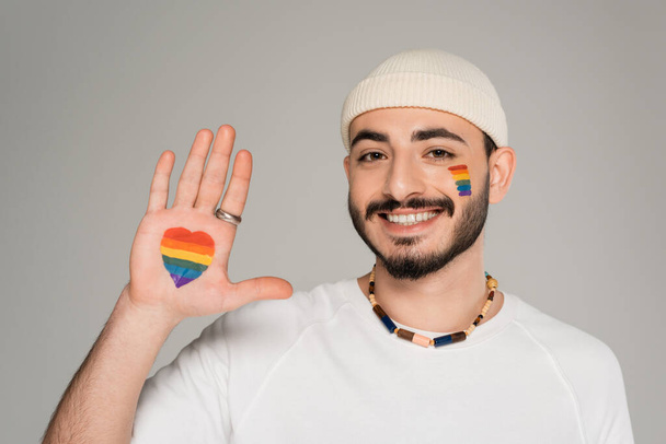Hombre homosexual sonriente con signo de corazón de la bandera lgbt en la mano aislado en gris   - Foto, imagen