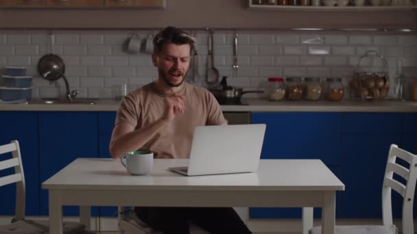 Egy fiatalember egy csésze kávéval leül az asztalhoz, és elkezd dolgozni egy laptopon. Egy férfi távolról dolgozik egy laptopon, miközben otthon ül a konyhában. Kiváló minőségű 4k felvételek - Felvétel, videó