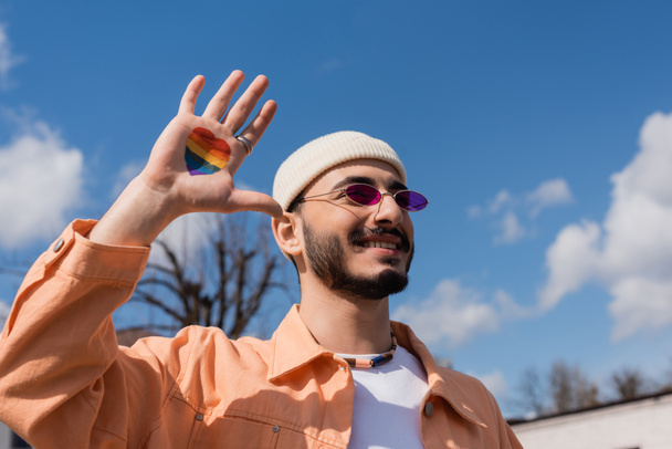 alegre gay hombre en gafas de sol con lgbt bandera en forma de corazón en la mano de pie al aire libre  - Foto, imagen