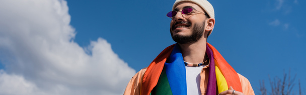 Vista de ángulo bajo del hombre gay despreocupado en gafas de sol con bandera lgbt de pie en la calle, pancarta  - Foto, imagen