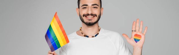 Fröhlicher homosexueller Mann mit lgbt-Fahne und Herz an der Hand isoliert auf grau, Banner  - Foto, Bild