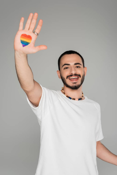 Carefree gay man showing lgbt flag in heart shape on hand isolated on grey, International day against homophobia - Photo, Image