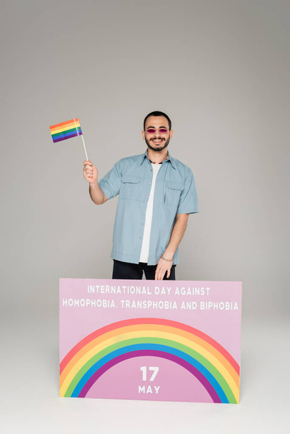 Alegre hombre gay sosteniendo bandera lgbt cerca de pancarta con Día Internacional contra la Homofobia, Transfobia y Bifobia letras sobre fondo gris  - Foto, Imagen