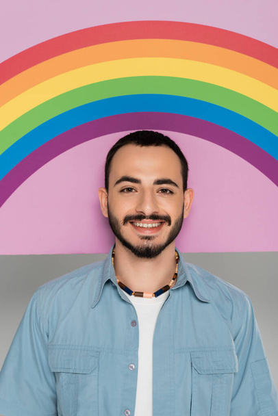 Portrait of smiling gay man looking at camera near placard with lgbt flag isolated on grey   - Photo, Image