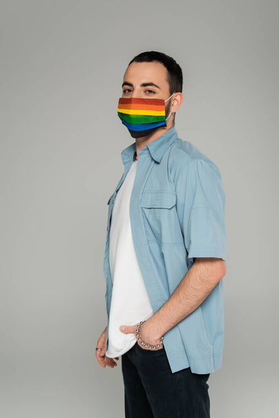 Morena gay hombre en médico máscara con lgbt bandera mirando cámara aislado en gris   - Foto, imagen