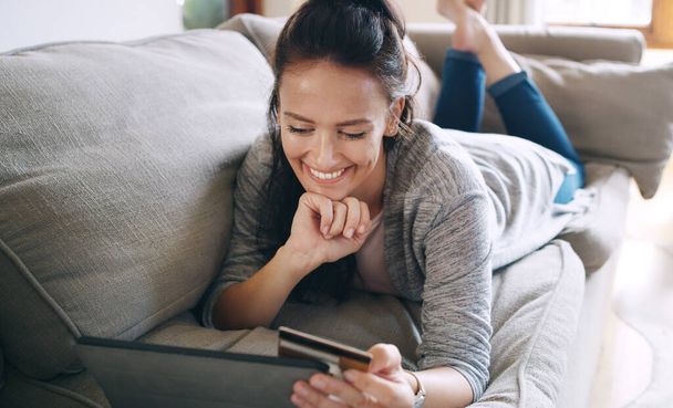 Looks like I bumped into a great discount. an attractive young woman using a digital tablet and a credit card to shop online while lying on a couch at home - Foto, Imagem