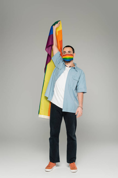 Full length of young gay man in medical mask holding lgbt flag on grey background  - Photo, Image