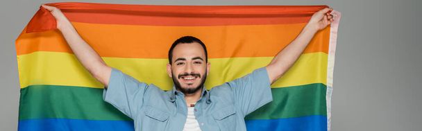 Ξέγνοιαστος ομοφυλόφιλος με σημαία lgbt και κοιτάζοντας την κάμερα απομονωμένη σε γκρι, banner  - Φωτογραφία, εικόνα