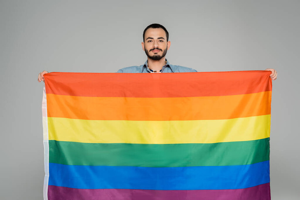 Hombre homosexual barbudo sosteniendo bandera lgbt y mirando a la cámara aislada en gris   - Foto, Imagen