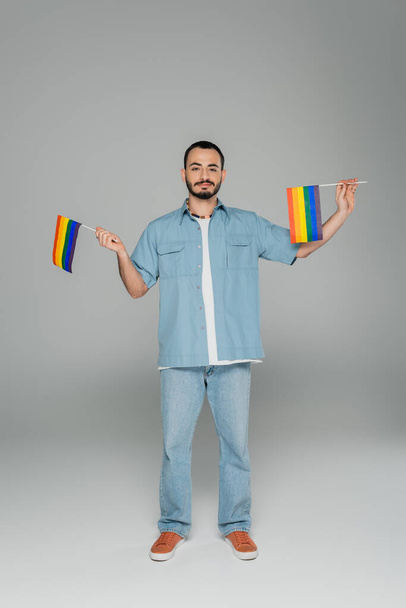 Full length of young gay man holding lgbt flags while standing on grey background, International Day Against Homophobia  - Photo, Image