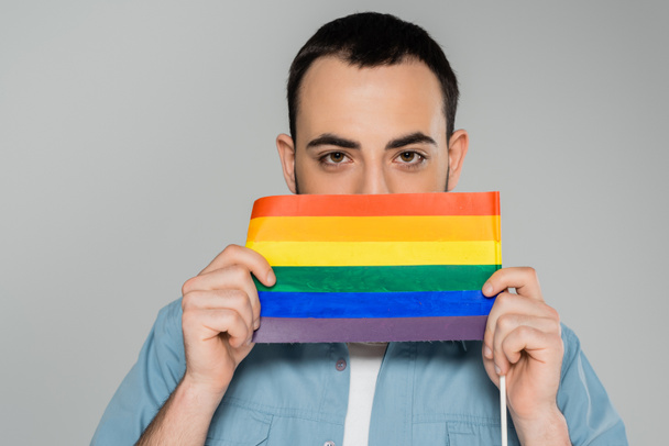Fiatal barna meleg férfi fedi arcát lgbt zászló elszigetelt szürke, Nemzetközi Homofóbia Elleni Nap  - Fotó, kép