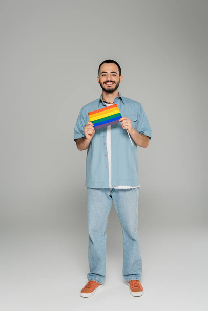 Full length of smiling homosexual man in denim shirt holding lgbt flag on grey background  - Photo, Image