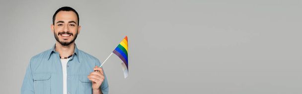 Μπρουνέτ γκέι άντρας χαμογελάει στην κάμερα και κρατάει την σημαία του lgbt απομονωμένη στο γκρι, banner  - Φωτογραφία, εικόνα