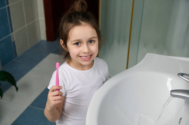 Χαρούμενο λευκό κοριτσάκι, που κρατάει οδοντόβουρτσα, βουρτσίζει δόντια, καθαρίζει το στόμα. Υγιής ανάπτυξη ευεξίας και φρέσκια αναπνοή. Πρωινή καθημερινή ρουτίνα. Οδοντιατρική περίθαλψη και στοματική υγιεινή - Φωτογραφία, εικόνα