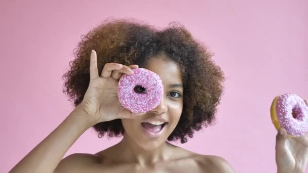 Linda menina adolescente afro-americana segurando donuts perto dos olhos com as mãos, modelo encaracolado com características faciais perfeitas, conceito de desnutrição, emoção pisca os olhos e sorri no fundo lilás rosa, lento - Filmagem, Vídeo