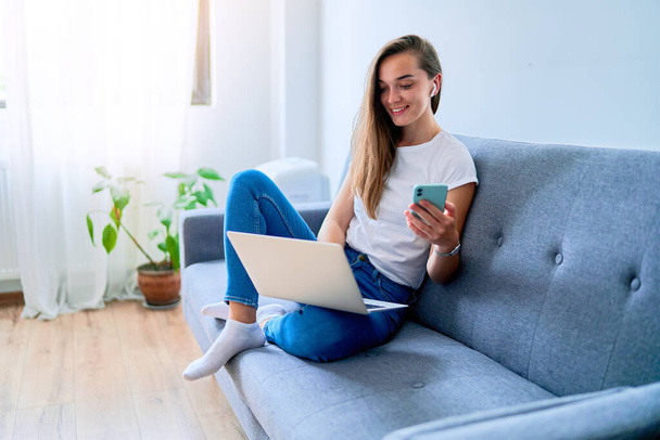 Young happy smiling joyful millennial girl blogger wearing wireless headphones sitting on a sofa and using laptop and phone for online remote work and browsing web at home  - Foto, immagini