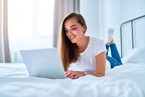 Young happy smiling attractive cute joyful millennial girl lying on a bed and using laptop for watching video online, learning language courses, social networking and video calling at home  - 写真・画像