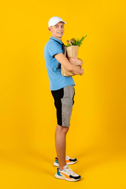 Young man in a blue t-shirt and cap holds a paper bag with food. Isolated on a yellow background. Delivery concept. - Photo, Image