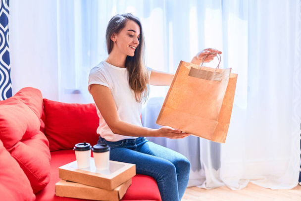Smiling cute satisfied happy casual adult joyful young millennial woman buyer received cardboard bags with takeaway food and drinks at home. Fast delivery service concept - Photo, Image