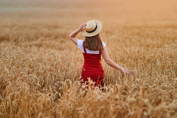 Back of calm serene free young female wearing straw hat and red denim overall standing in golden yellow dry wheat field and enjoying beautiful freedom moment  - Foto, imagen