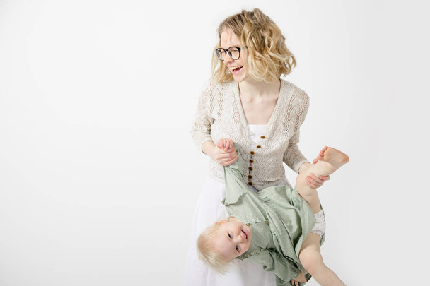 Porträt einer glücklichen Familie, die auf weißem Hintergrund posiert. Junge fröhliche attraktive Frau Mutter hält Bein, Hand von kleinen blonden Mädchen Tochter trägt grünes Kleid, Spaß haben, spielen. Kindheit. - Foto, Bild