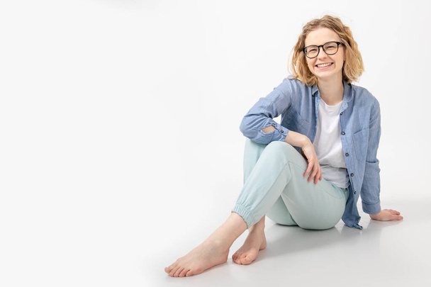 Portrait of young cheerful barefoot woman with short wavy hair wearing glasses, denim shirt, blue trousers sitting with crossed legs, looking at camera, leaning on hand on white background. Studio. - Foto, imagen
