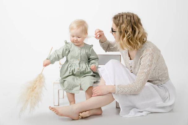 Portrait of cute little blond girl wearing green dress, sitting on white little bench on white background, holding branch of dried pampas grass. Young woman tickling face with twig. Childhood, family. - Photo, Image