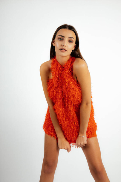 Portrait of young sensual woman with long dark hair, natural make-up wearing short fluffy orange halter dress, standing, bending forward on white background. Beauty, fashion, vogue. Vertical, studio. - Фото, изображение