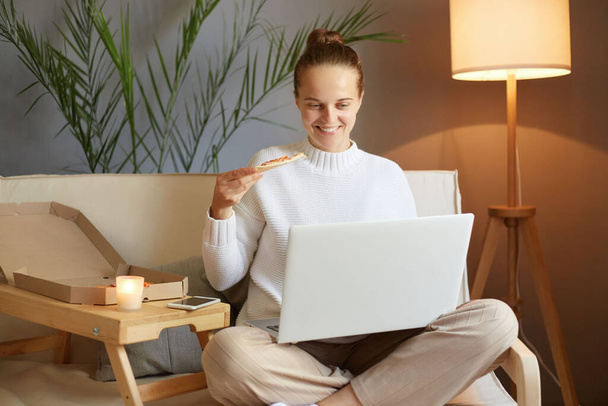 Relaxed happy smiling woman with bun hairstyle in white sweater looking at laptop screen and having snack eating pizza while watching film sitting on sofa at home. - Photo, Image