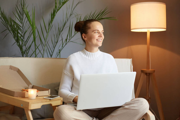 Dreamy smiling caucasian woman with bun hairstyle in white sweater looking at laptop screen sitting on sofa at home looking away with happy expression enjoying her remote work. - Фото, изображение