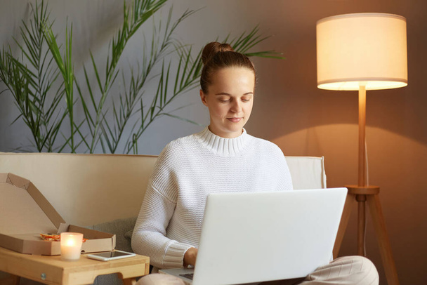 Remote work. Freelance job. Concentrated focused woman with bun hairstyle in white sweater looking at laptop screen typing keyboard working sitting on sofa at home. - Foto, Bild