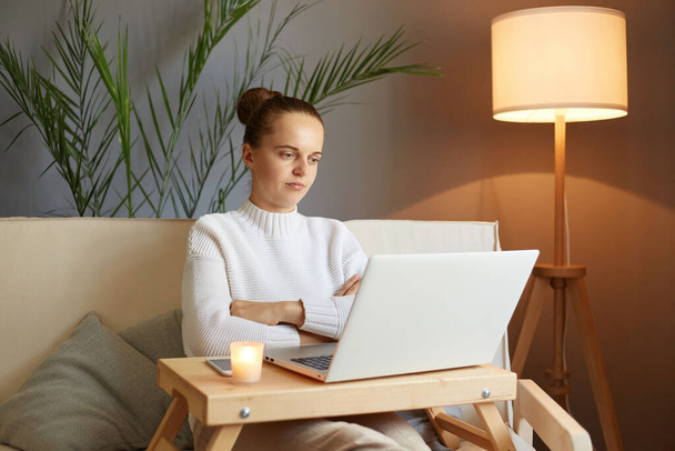 Bored sad woman with bun hairstyle in white sweater looking at laptop screen with sorrowful expression watching uninteresting video or having boring video call sitting on sofa at home - Foto, Imagen