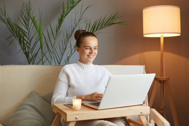 Optimistic joyful positive woman with bun hairstyle in white sweater looking at laptop screen sitting on sofa at home having online conference working online or watching content in social network. - Foto, afbeelding