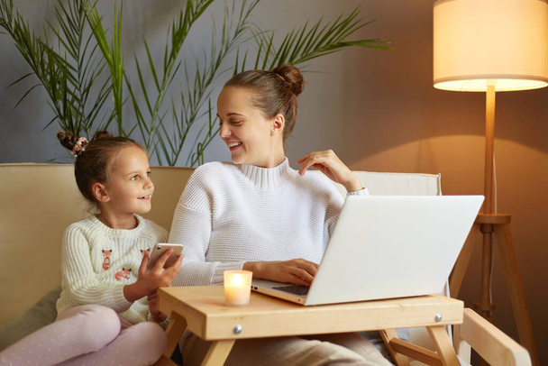 Smiling woman with her little kid posing at home mother with daughter watching movie together on laptop while sitting on sofa enjoying leisure time. - Photo, Image